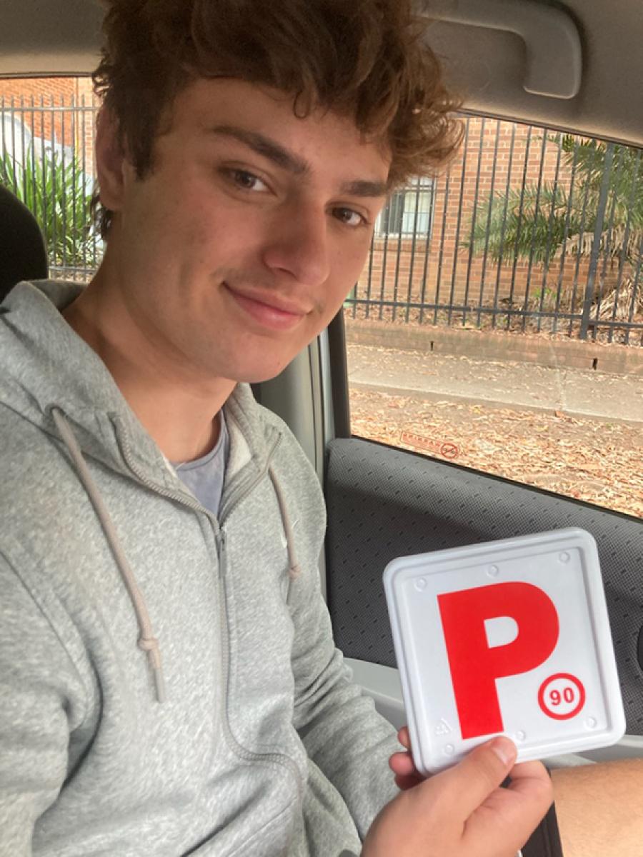 Smiling learner driver in a car holding a red P plate after driving lessons