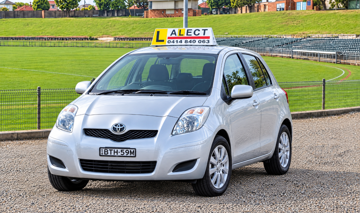 White Toyota Yaris learner driver car with roof sign parked beside a green playing field