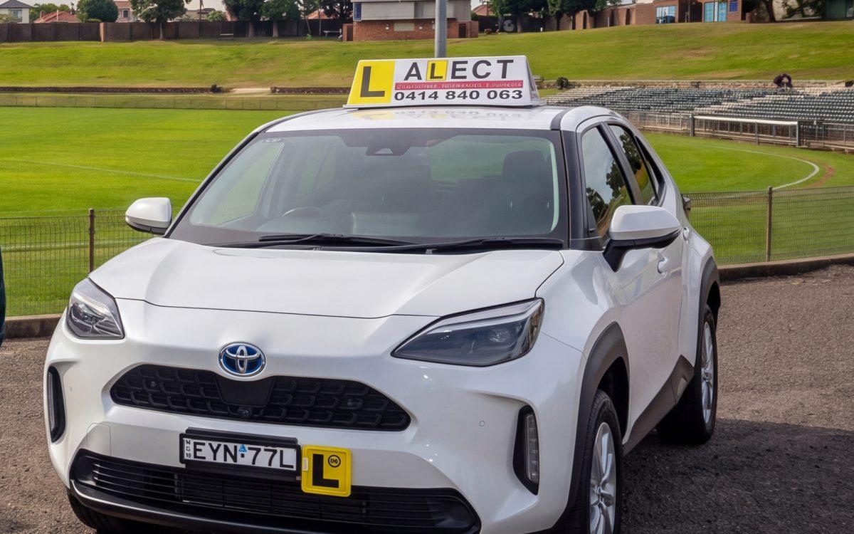 White learner driver car with roof sign parked near a green public space